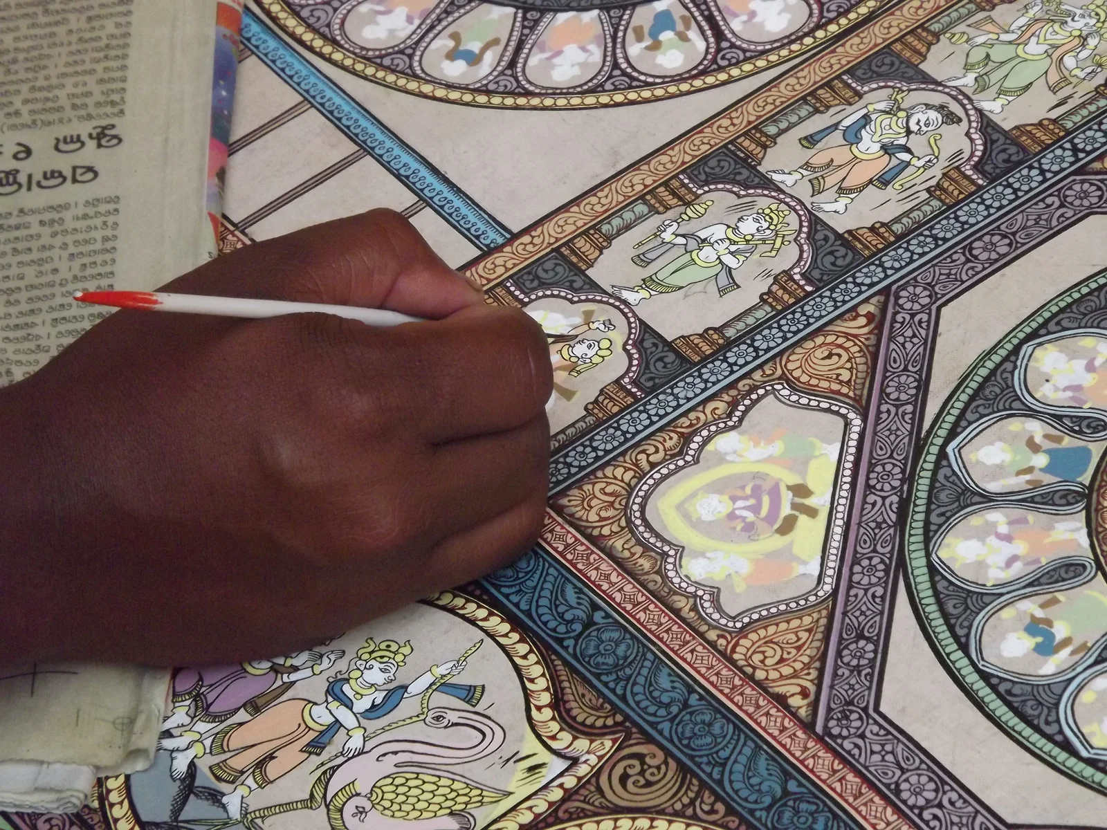 An artist’s hand uses a fine brush on a Pattachitra with figures and patterned borders.
