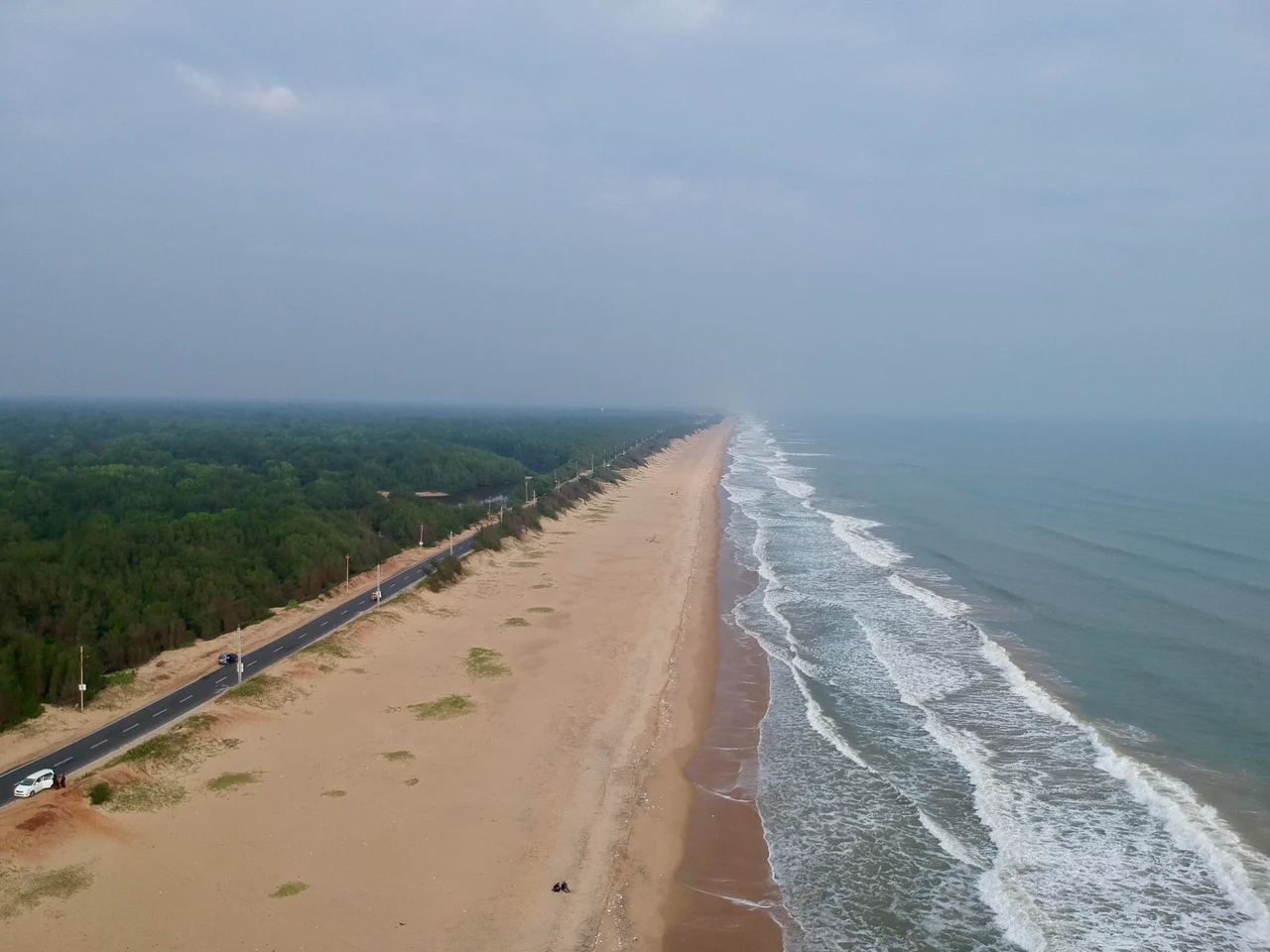 An aerial view of a long sandy shore, surf, a coastal road and trees at Chandrabhaga.