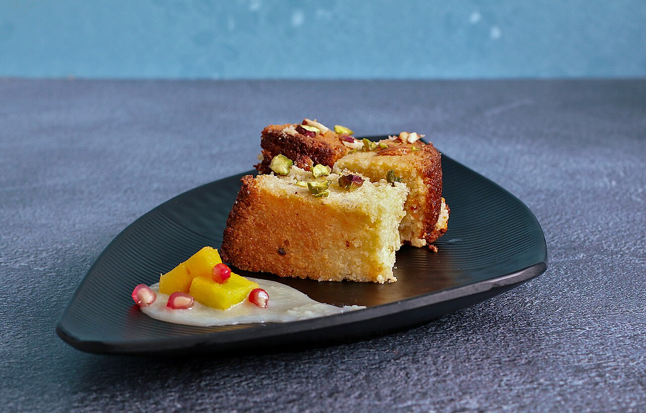 Two golden pieces of chhena poda on a dark plate, with fruit and a garnish.