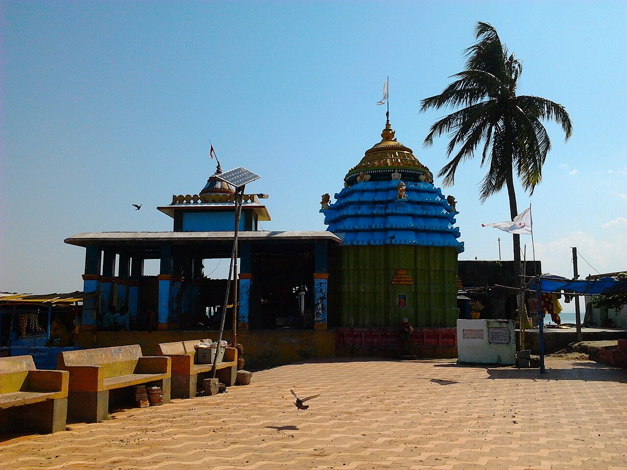 The blue Kalijai temple beside a palm tree, with its courtyard in the foreground.