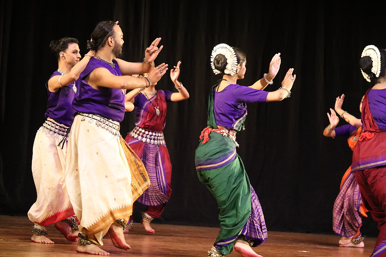 A group of Odissi dancers performing on stage in purple, green and cream costumes.