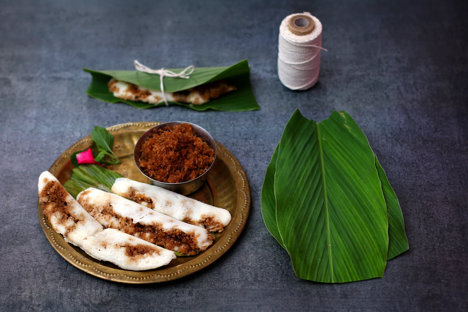 Opened Enduri pitha with brown filling on a brass-coloured plate, beside green leaves and a wrapped parcel.