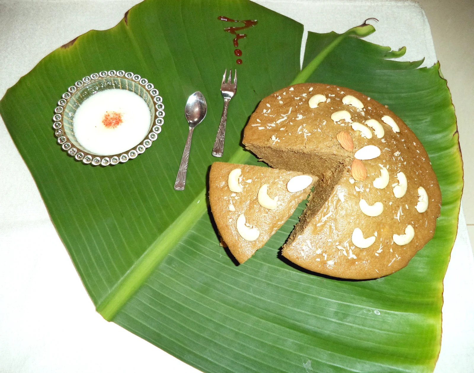 Round Poda pitha with a cut slice and nut garnish, presented on a green leaf beside a small bowl.