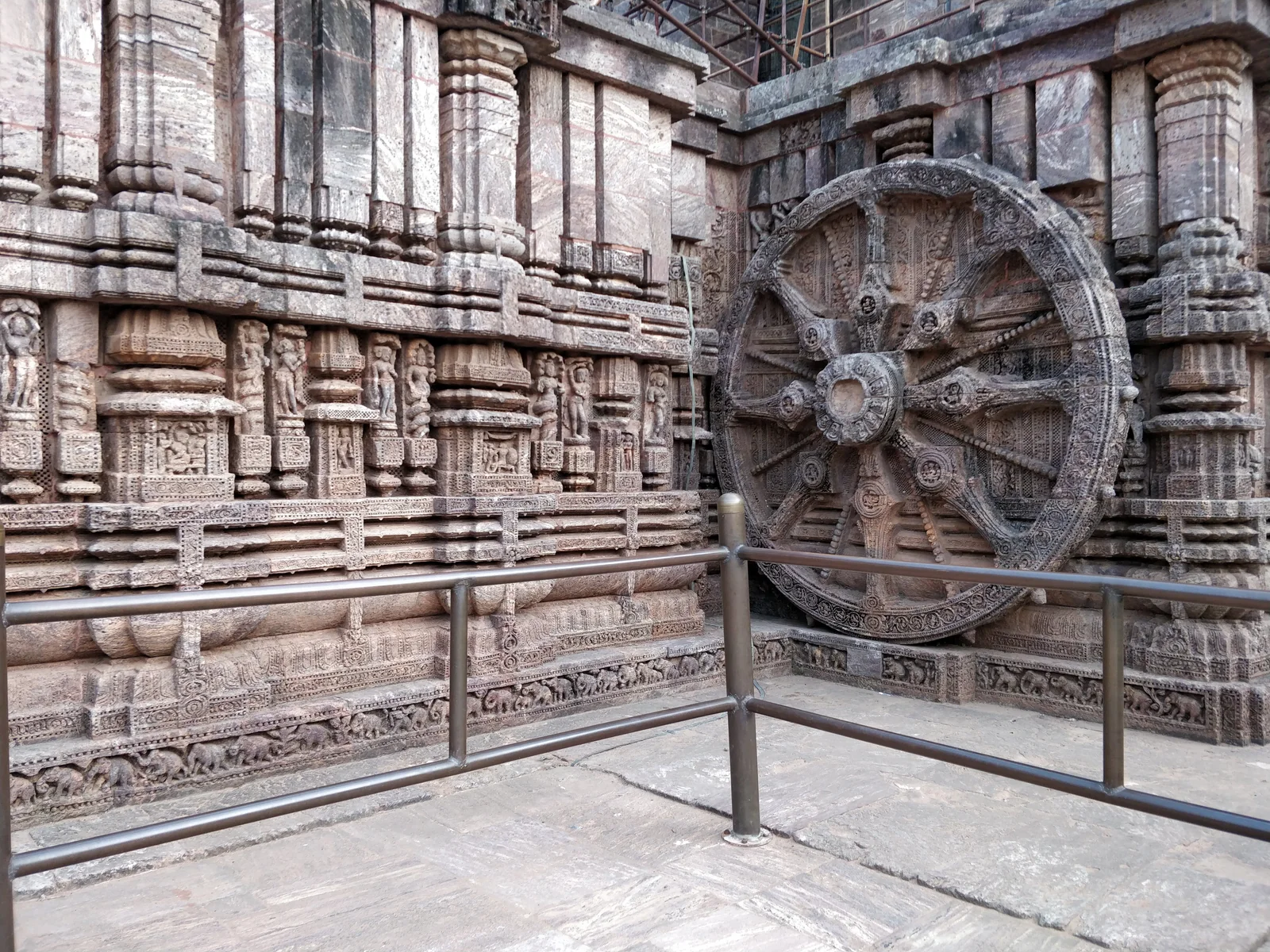 A richly carved stone chariot wheel and relief-covered wall at Konark Sun Temple, with protective metal railings in front.
