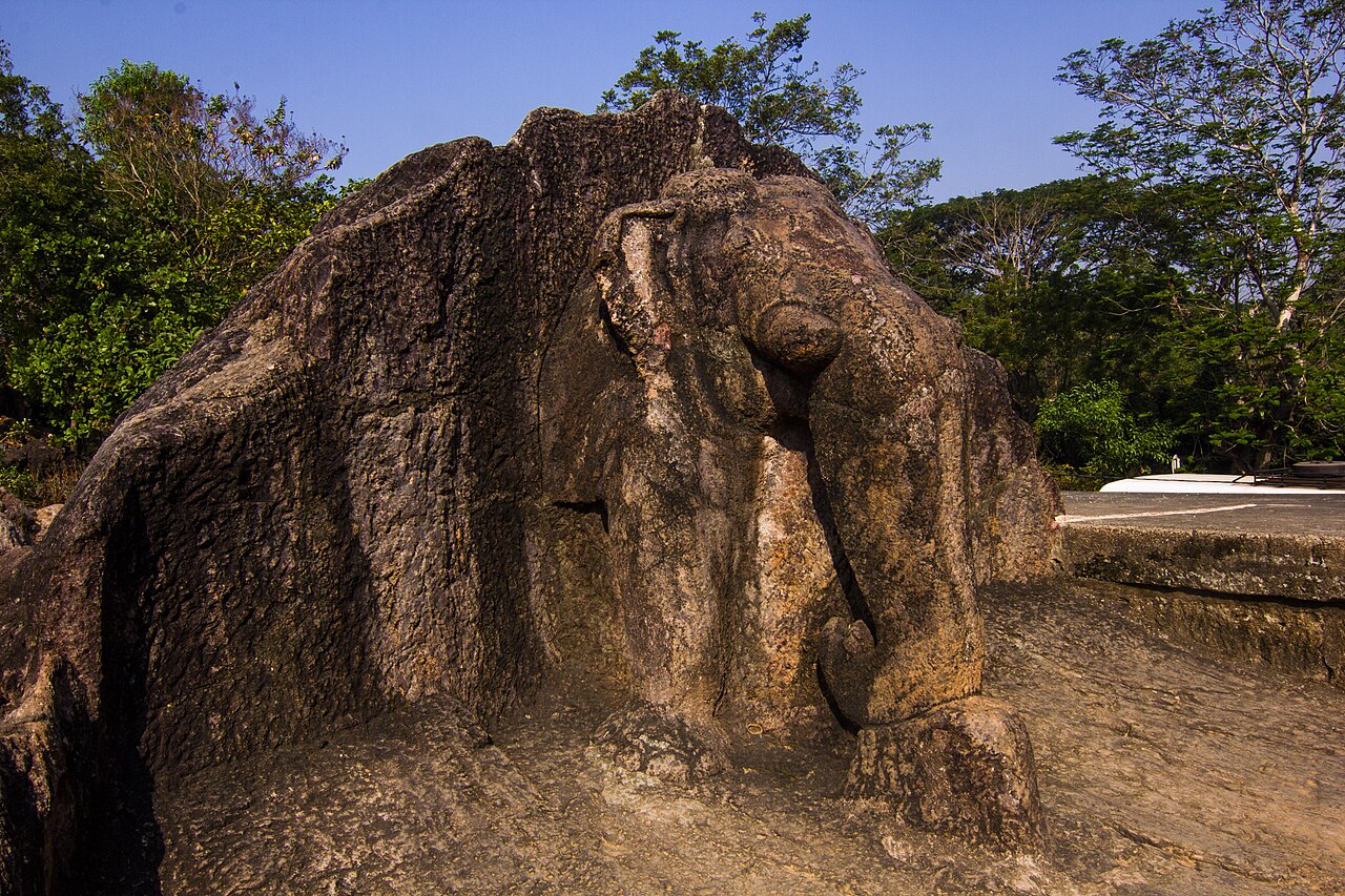 The front of an elephant carved from a large rock, with trees behind it.