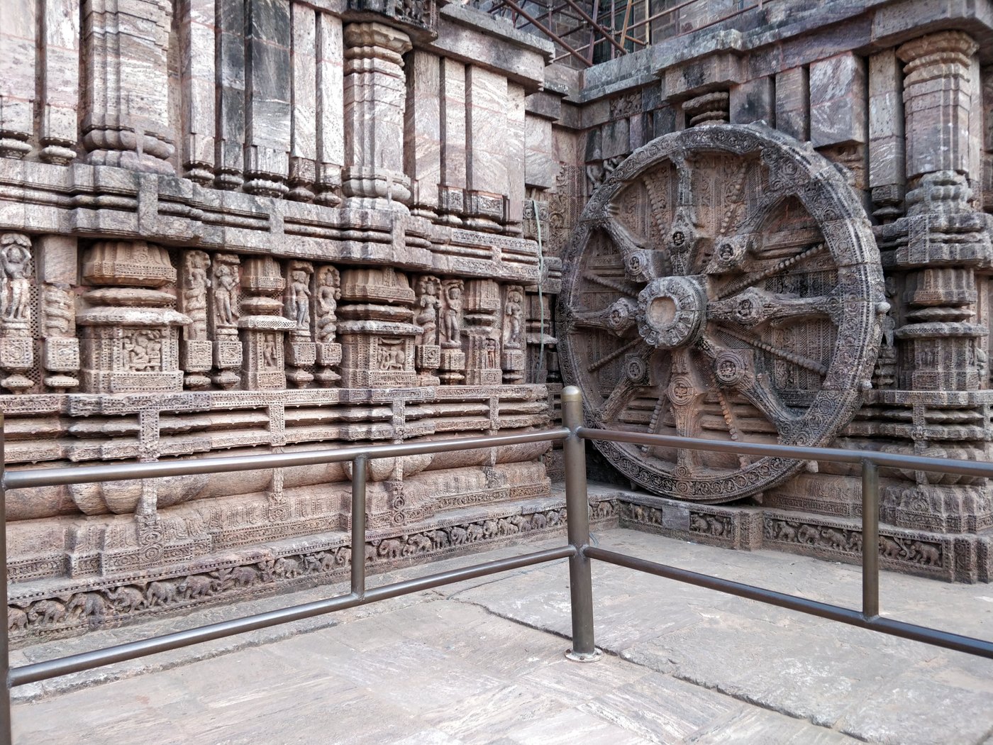 A richly carved stone chariot wheel and relief-covered wall at Konark Sun Temple, with protective metal railings in front.
