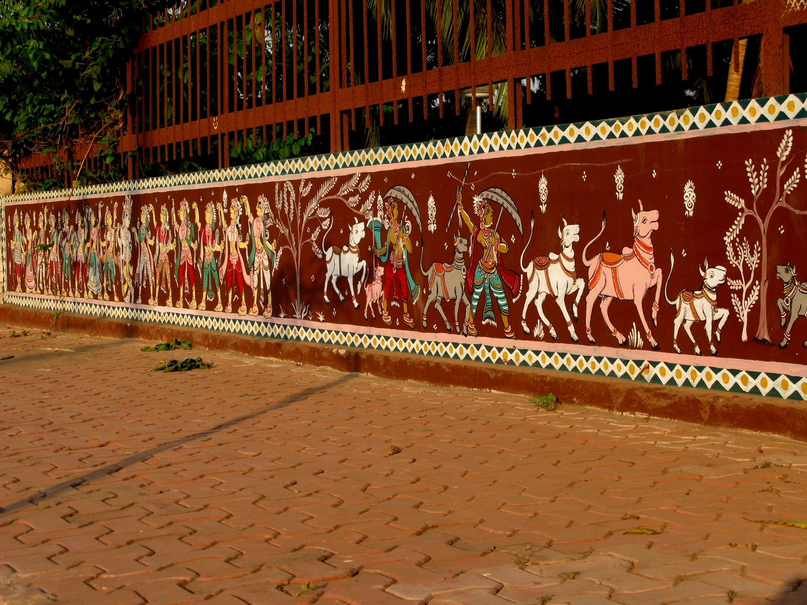 A long red wall painted with a procession, beside a brick pavement.
